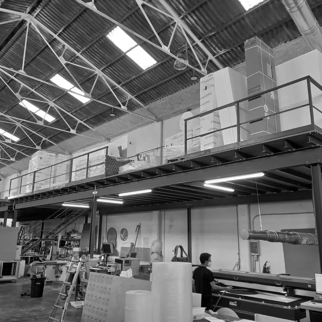 Interior de almacén con entrepiso de acero, mostrando áreas de trabajo debajo y opciones de almacenamiento en el nivel superior.